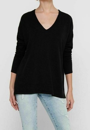 Frau trägt ein locker sitzendes schwarzes Langarm-V-Neck-Oberteil und hellblaue, abgenutzte Jeans vor einem schlichten hellen Hintergrund.