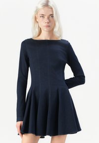 Marineblauwe lange jurk met lange mouwen, een getailleerde lijfje en een uitlopende rok, gemaakt van een soepele stof. Heeft verticale naden en een bootheksboord.