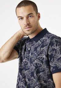 Marineblauw poloshirt met een wit tropisch bladpatroon, korte mouwen, een puntige kraag en een klein logo op de borst.
