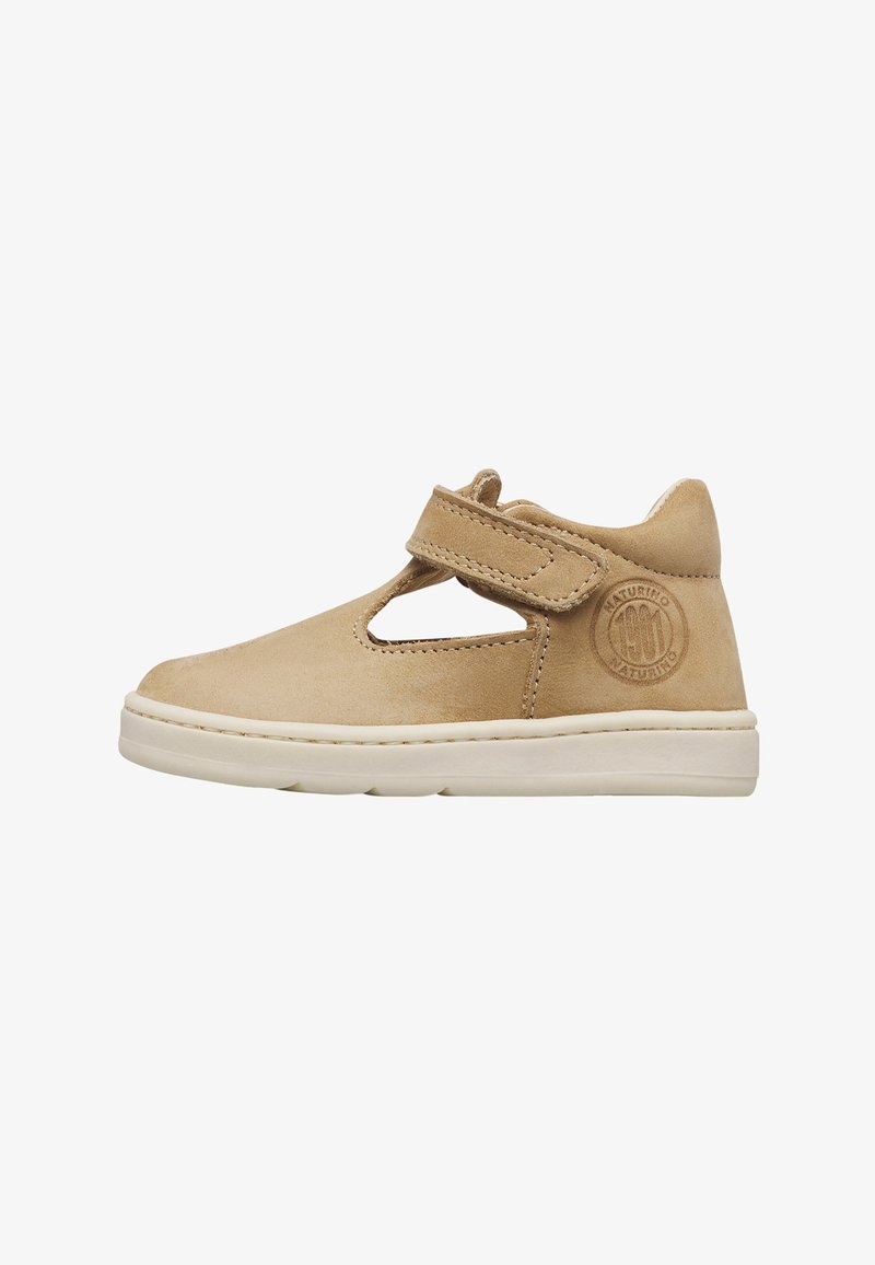 Chaussure enfant beige en suède avec découpe latérale, bride à scratch, semelle blanche et logo de marque circulaire en relief près du talon.