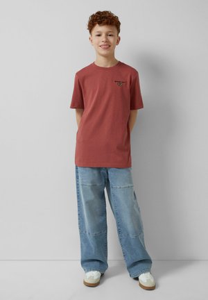 T-shirt z bawełny w kolorze rdzy, z okrągłym dekoltem i krótkimi rękawami, z małym czarnym nadrukiem na lewej piersi. W zestawieniu z luźnymi niebieskimi dżinsami.