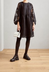 Robe noire avec manches transparentes bouffantes et taille froncée. Associée à des collants noirs et des mocassins marron épais. Tenant un petit sac à main blanc.