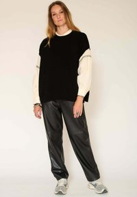 Un gros pull noir surdimensionné superposé à une blouse crème à manches bouffantes, associé à un pantalon en simili cuir noir et des baskets grises.