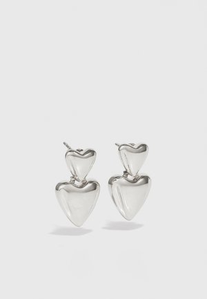 Pendientes de plata en forma de corazón, presentando un corazón más grande debajo de uno más pequeño. Superficie lisa y reflectante con un acabado pulido.