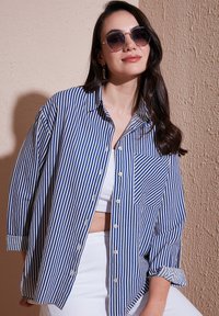 Camicia a righe blu e bianche con taschino sul petto, vestibilità rilassata e maniche arrotolate. Indossata sopra una maglietta bianca e abbinata a occhiali da sole.