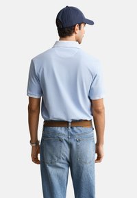 Mand iført lyseblå poloshirt, blå baseballkasket, blå jeans og brun bælte, vist bagfra på hvid baggrund.