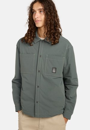 Jong persoon met lang krullend haar, draagt een grijze knopjesjas met twee zakken aan de voorkant en beige broek, handen in de zakken.