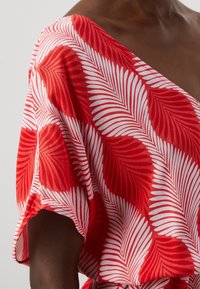 Haut à motifs rouges et blancs présentant un design de feuilles de palmier superposées. Tissu doux et léger avec une coupe ample et des manches larges.