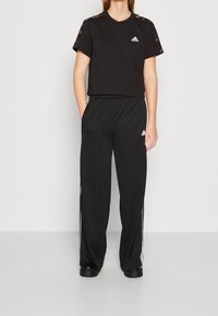 Černé krátké tričko s výřezovými detaily na ramenou a bílým logem. Černé kalhoty s elastickým pasem a bílými pruhy po stranách.