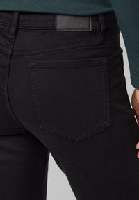 Gros plan sur la poche arrière d'un jean en denim noir et les passants de ceinture, porté avec un haut vert foncé, montrant les coutures et la patch en cuir de la marque.