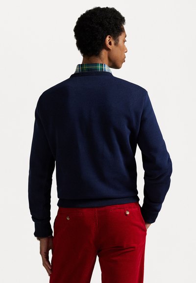 Marineblauer Baumwollpullover mit strukturiertem Rücken, darunter ein kariertes Hemd mit Kragen, kombiniert mit roten Cordhosen.