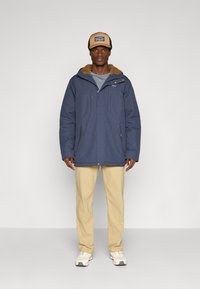 Parka bleumarin cu glugă căptușită cu blană, două buzunare pe piept și două buzunare laterale, purtată peste o cămașă gri și asortată cu pantaloni bej.