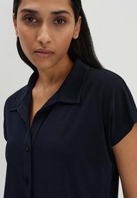 Camicetta corta blu navy con colletto, dotata di chiusura a bottoni e una texture liscia e morbida. Design semplice e strutturato.