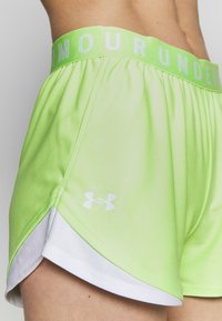 Zbliżenie na osobę noszącą szorty sportowe Under Armour w limonkowym kolorze z białymi wykończeniami i elastycznym pasem z logo.