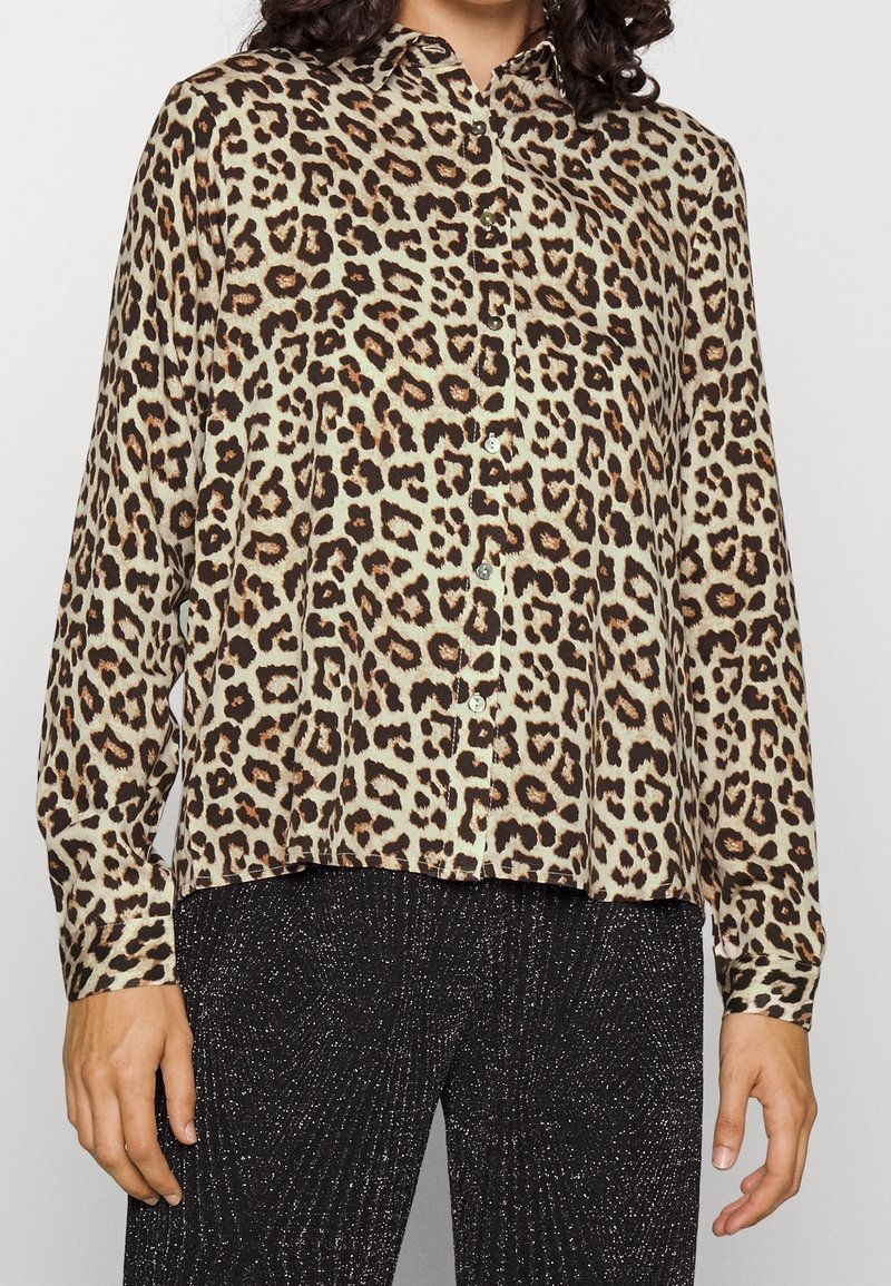 Femme portant une chemise boutonnée à manches longues imprimé léopard et un pantalon noir pailleté, debout les bras détendus le long du corps.