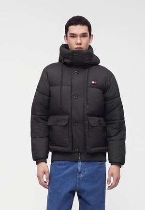 Junger Mann trägt eine schwarze Steppjacke mit hohem Kragen und blaue Jeans, steht vor einem einfarbigen hellen Hintergrund.