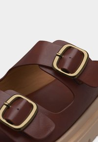 Sandale en cuir marron avec deux larges brides ornées de boucles rectangulaires dorées et une semelle beige rembourrée.