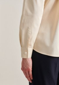 Light beige long-sleeve shirt with a buttoned cuff and a tailored fit. Smooth texture, subtle sheen, and no visible patterns.