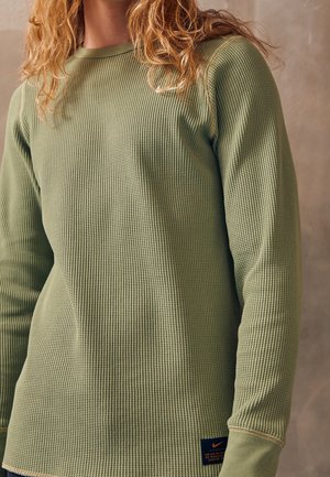 Person trägt ein langärmliges, olivgrünes Waffelstrickshirt mit einem kleinen Nike-Logo-Patch nahe dem Saum.