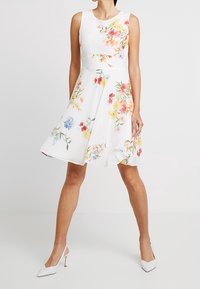 Abito bianco senza maniche con un motivo floreale in rosso, giallo e blu, silhouette A-line, con un corpetto aderente e una gonna svasata.