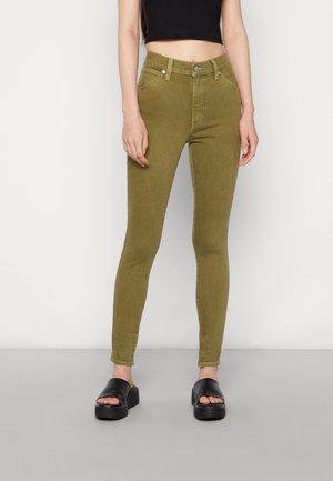 Jeans skinny verde oliva con cintura alta, bolsillos frontales y cierre de cremallera. Llevados con sandalias negras sin hebilla. Tejido liso y elástico.