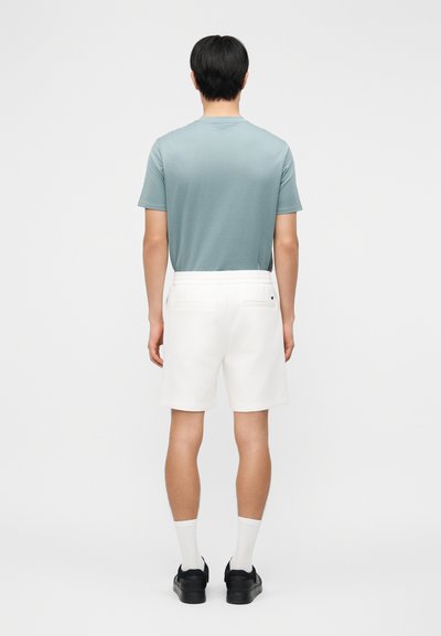 Άνδρας στέκεται πλάτη, φοράει γαλάζιο t-shirt, λευκά σορτς, λευκές κάλτσες και μαύρα αθλητικά παπούτσια σε απλό φόντο.