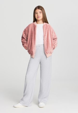 Giacca bomber in peluche rosa con texture a costine, abbinata a pantaloni di felpa a gamba larga grigio chiaro. Le sneakers bianche completano l'outfit.