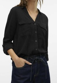 Camicia nera con colletto, due tasche sul petto e maniche arrotolate. Indossata con jeans blu scuri a vita alta.