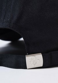 Gorra negra con una tela texturizada, que presenta un cierre metálico plateado con la firma en relieve. La correa es ajustable y trenzada.