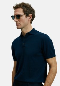 Polo shirt bleu marine à manches courtes, avec un plastron à deux boutons, fabriqué à partir d'un tissu lisse. Accompagné de lunettes de soleil sombres, le modèle se tient de profil à gauche.