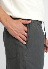Grijze broek van een gestructureerde stof heeft een ritszak aan de zijkant. Een hand steekt in de zak, waarbij een zilveren ring op een vinger zichtbaar is.