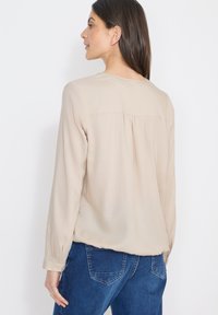 Blouse beige clair à manches longues, texture douce, détail de plissage à l'arrière, complétée par un col rond. Associée à un jean bleu.