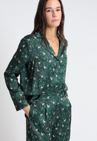Zelená saténová pyžamová souprava s vzorem jelenů a stromů. Obsahuje límeček, knoflíky a kapsu. Hladký materiál a přiléhavý střih.