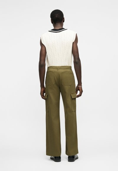 Άνδρας που φοράει λευκό αμάνικο γιλέκο πλεκτό με μαύρες λεπτομέρειες και ελαι πράσινο παντελόνι cargo, όρθιος με την πλάτη προς το μέρος μας, σε απλό φόντο