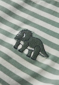 Grün-weiß gestreuter Stoff mit einem bestickten Dinosaurierdesign in dunklerem Grün, das strukturierte Stiche und kreisförmige Akzente zeigt.