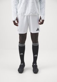 Hvid sportsjersey med abstrakt mønster, hvide shorts med logo, sorte sokker med hvide striber og sorte fodboldstøvler med struktureret overflade.