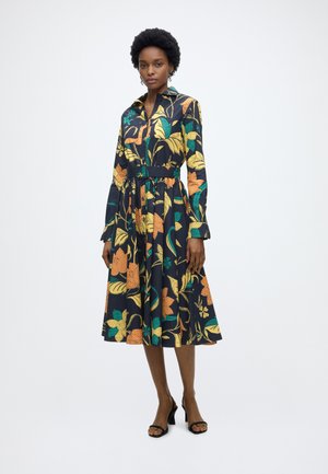 Frau steht in einem schwarzen, taillierten Midikleid mit langen Ärmeln und großem gelb-orange-grünem Blumenmuster, kombiniert mit schwarzen High Heels.