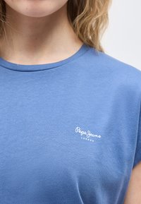 Κοντινό πλάνο ενός ατόμου που φοράει μπλε μπλουζάκι με στρογγυλή λαιμόκοψη και μικρό λευκό λογότυπο Pepe Jeans London στο στήθος.