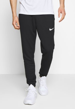 Mann, trägt schwarze Nike-Sweatpants und weiße Sneaker, steht vor einem schlichten weißen Hintergrund.
