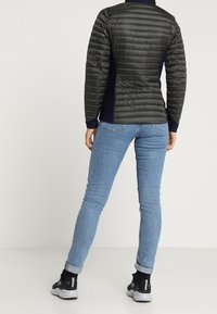 Svart och grön vadderad jacka med dragkedja i kragen, figurnära design, kombinerad med blå jeans och svarta träningssko. Slät textur, quiltat mönster.