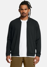 Schwarze Zip-Jacke mit einfachem Design, gerippten Bündchen und glatter Textur, getragen über einem weißen T-Shirt und kombiniert mit beigen Hosen.
