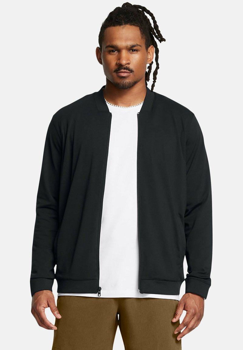 Schwarze Zip-Jacke mit einfachem Design, gerippten Bündchen und glatter Textur, getragen über einem weißen T-Shirt und kombiniert mit beigen Hosen.