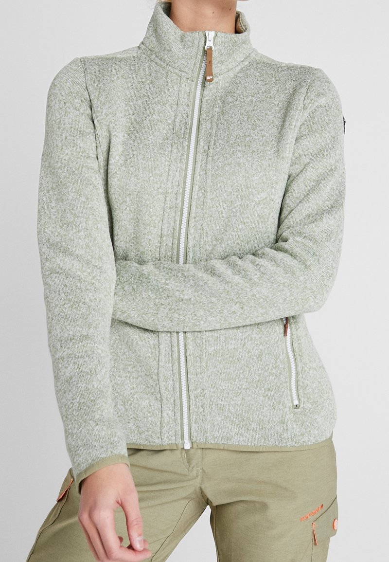 Personne portant une veste polaire zippée gris clair avec col montant et un pantalon cargo beige avec un logo orange.