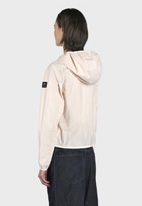 Leggera giacca a vento rosa pallido con cappuccio, polsini elasticizzati e una patch del logo nera sulla manica. Presenta una texture liscia e lucida.