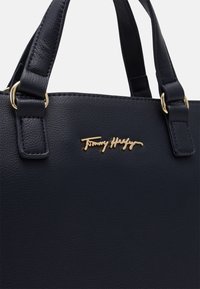 Czarna torebka skórzana z fakturą, z podwójnymi uchwytami i złotym logo Tommy Hilfiger umieszczonym na środku z przodu.