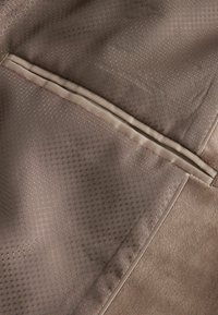 Brauner Stoff mit einem strukturierten Wabenmuster und einem glatten, kontrastierenden beigen Besatz entlang der Naht.