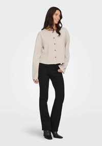 Jasno beżowy krótki kardigan z okrągłym dekoltem, wyraźnymi guzikami i szerokimi rękawami. Zestawiony z czarnymi dopasowanymi spodniami i botkami na obcasie.