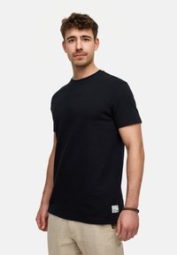 Camiseta negra de manga corta en tejido de punto suave que presenta un cuello redondo y una pequeña etiqueta cerca del dobladillo. Se lleva con pantalones de color claro.