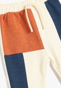 Pantalones de chándal con bloques de color en secciones naranja, crema y azul marino, hechos de tela suave con una cintura elástica y cordón.