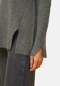 Grau gerippter Pullover mit seitlichen Schlitzen, langen Ärmeln und lockerer Passform. Das Material wirkt weich und strukturiert. Über einer dunklen Jeans getragen.